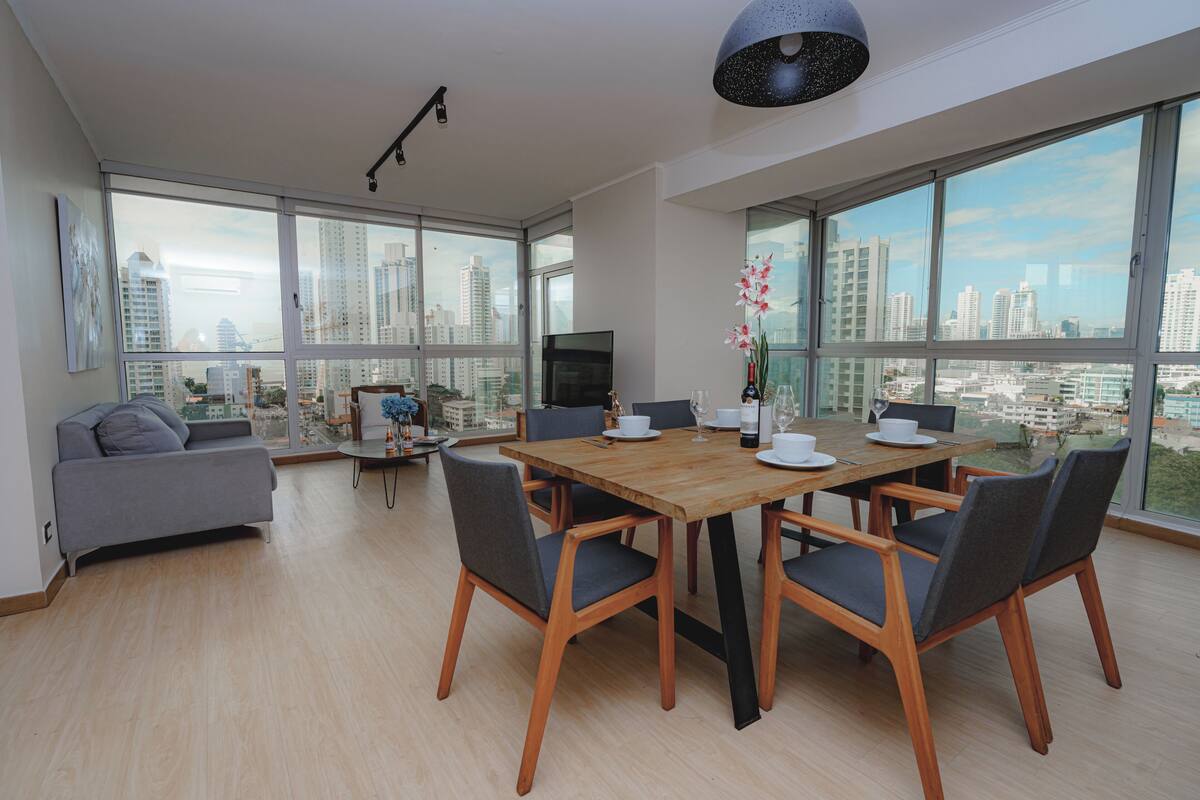 Ein moderner Essbereich mit einem Holztisch, grauen Stühlen und Blick auf die Stadt durch große Fenster.