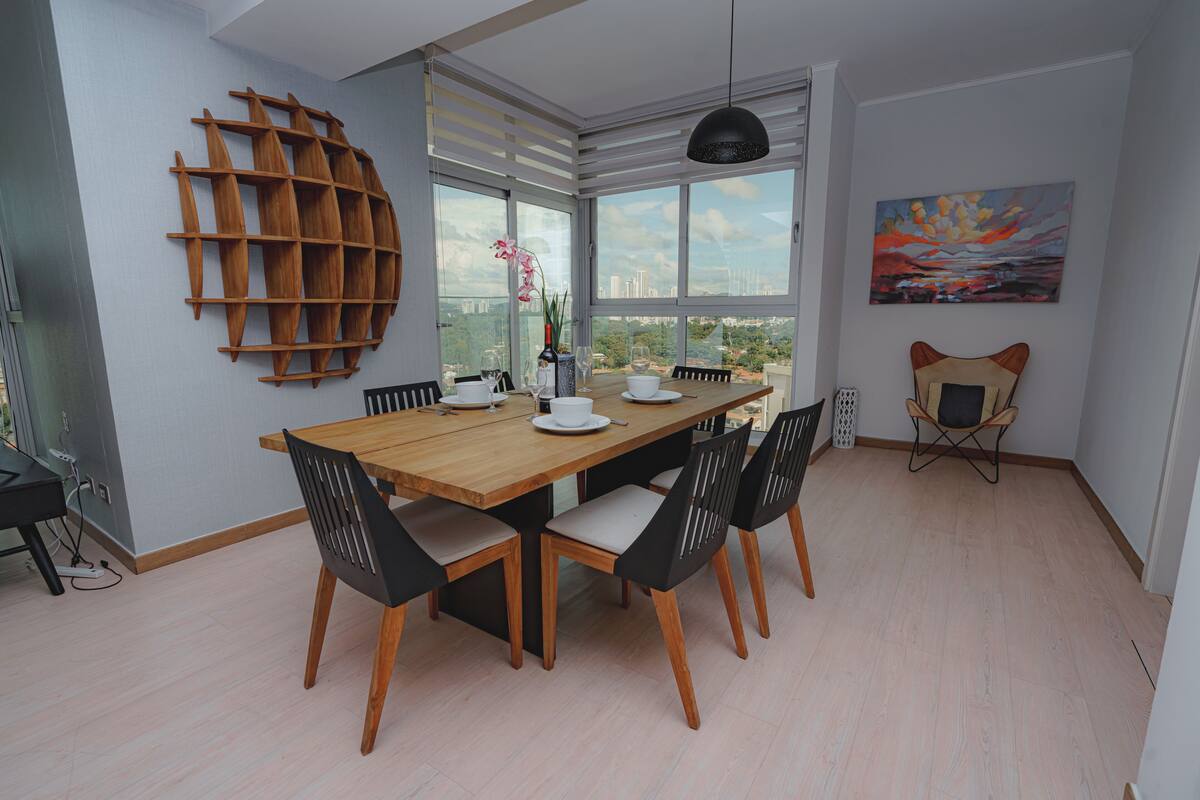 Ein Essbereich mit einem Holztisch, schwarzen Holzstühlen, einer schwarzen Pendelleuchte und Blick auf die Stadt durch große Fenster.