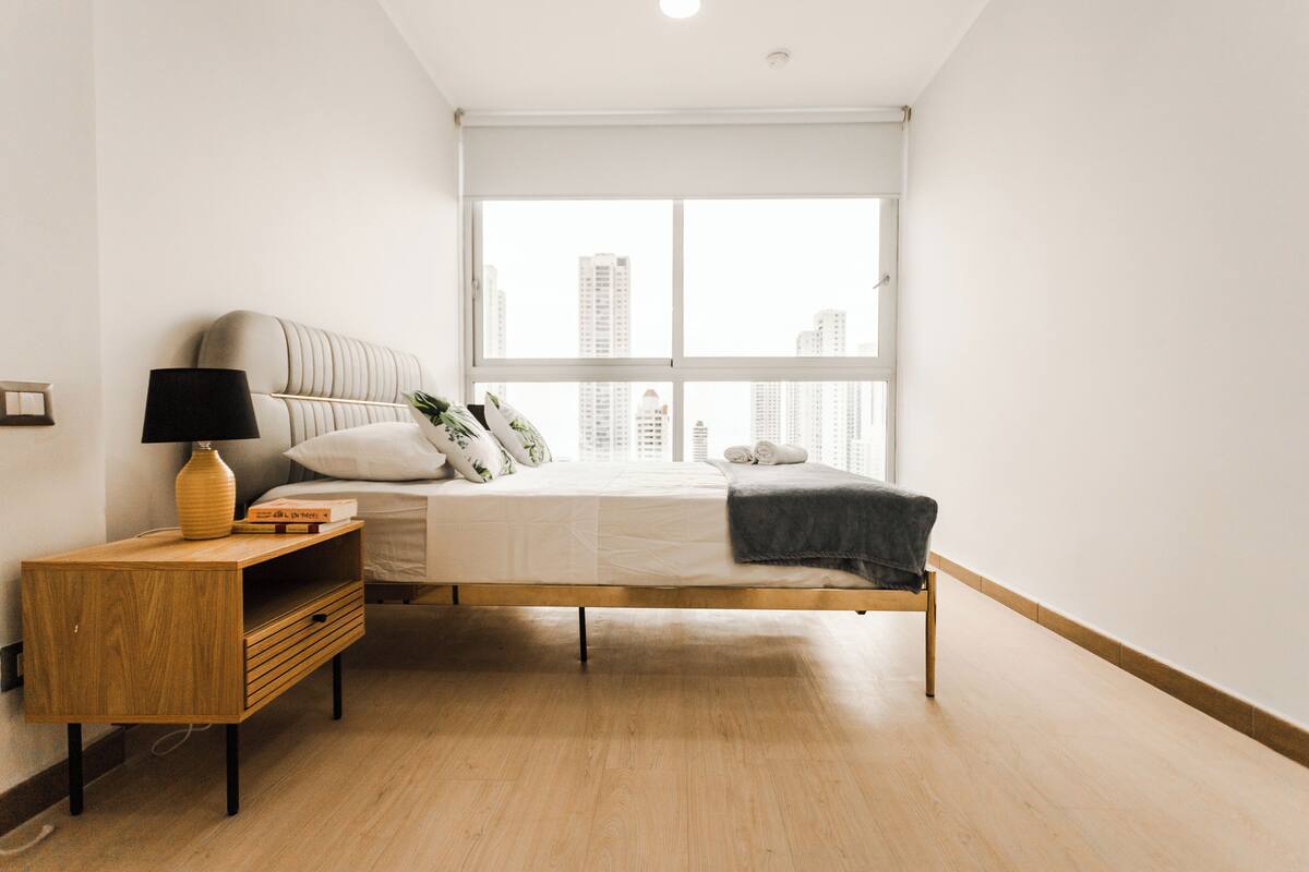 Ein schlicht eingerichtetes Schlafzimmer mit Bett, Nachttisch, Lampe und Blick auf hohe Gebäude durch ein großes Fenster.