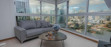 A modern living room with a grey sofa, a round coffee table, and large windows offering a city view.
