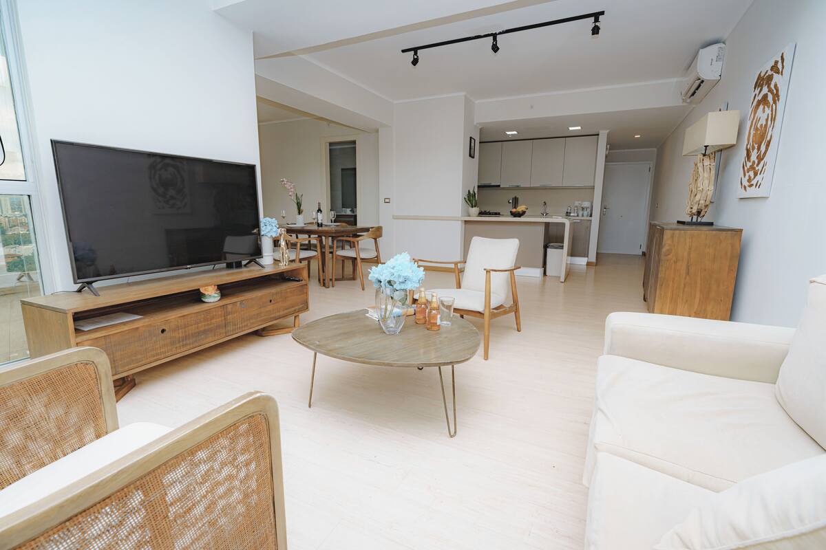 Ein Wohnzimmer mit Fernseher, rundem Couchtisch und einer Couch. Im Hintergrund befindet sich ein Essbereich mit Tisch und Stühlen.