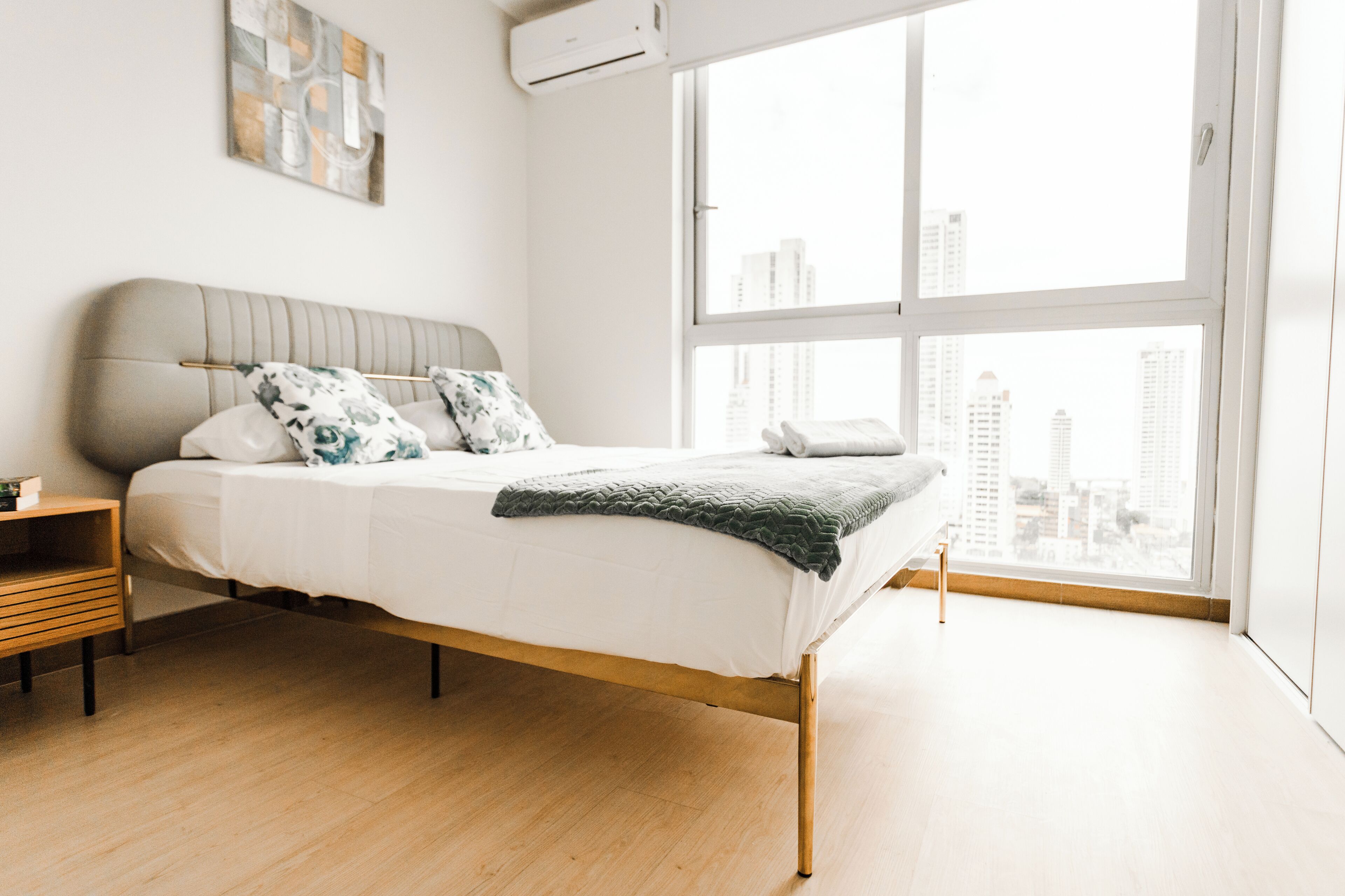 Un dormitorio moderno con un ventanal grande, una cama con cabecera gris y una mesita de noche de madera.