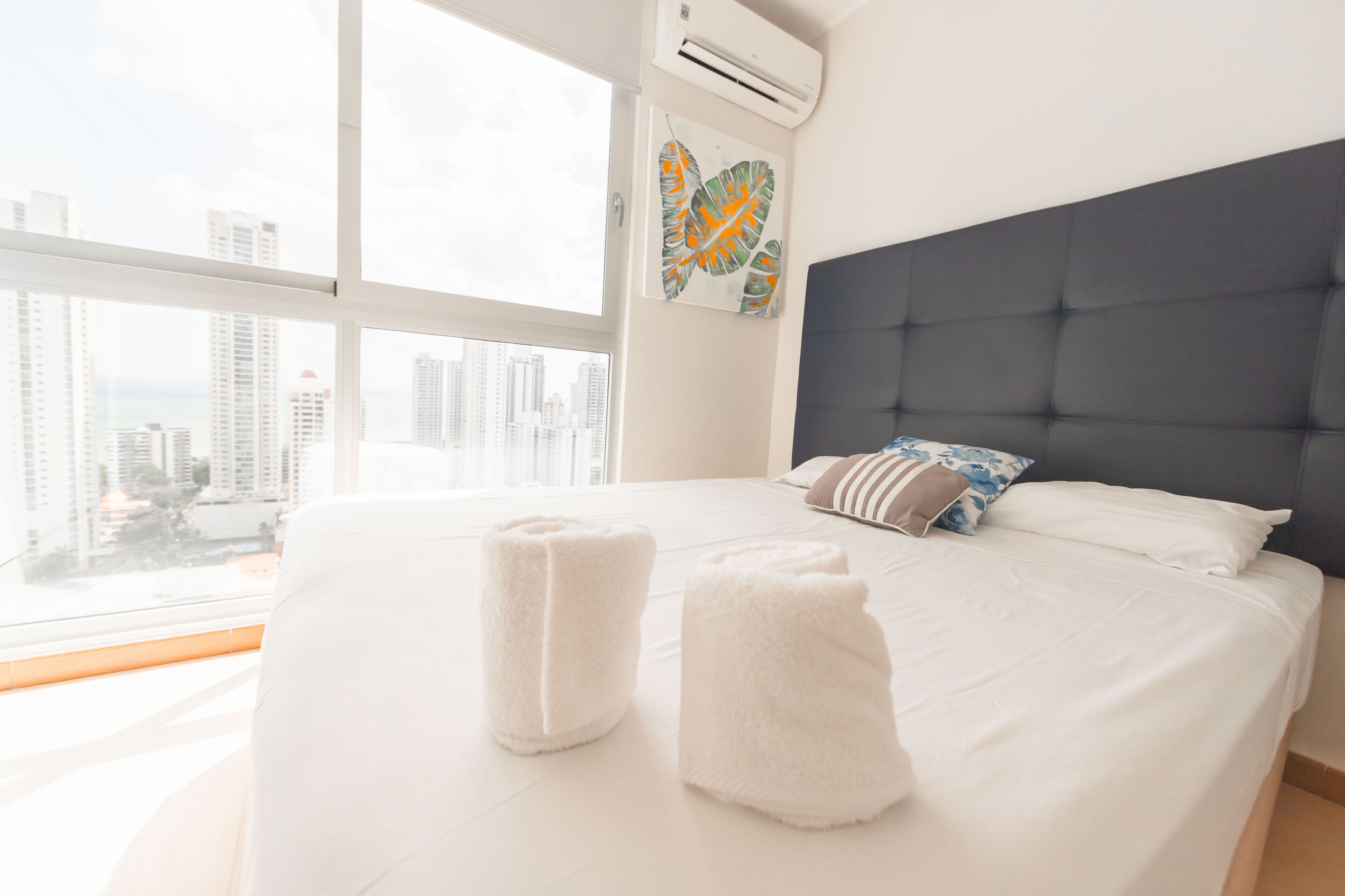 A hotel room with a large bed, white linens, a dark headboard, and a city view through large windows.