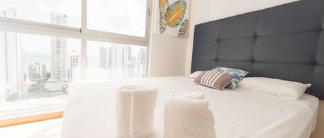 A hotel room with a large bed, white linens, a dark headboard, and a city view through large windows.