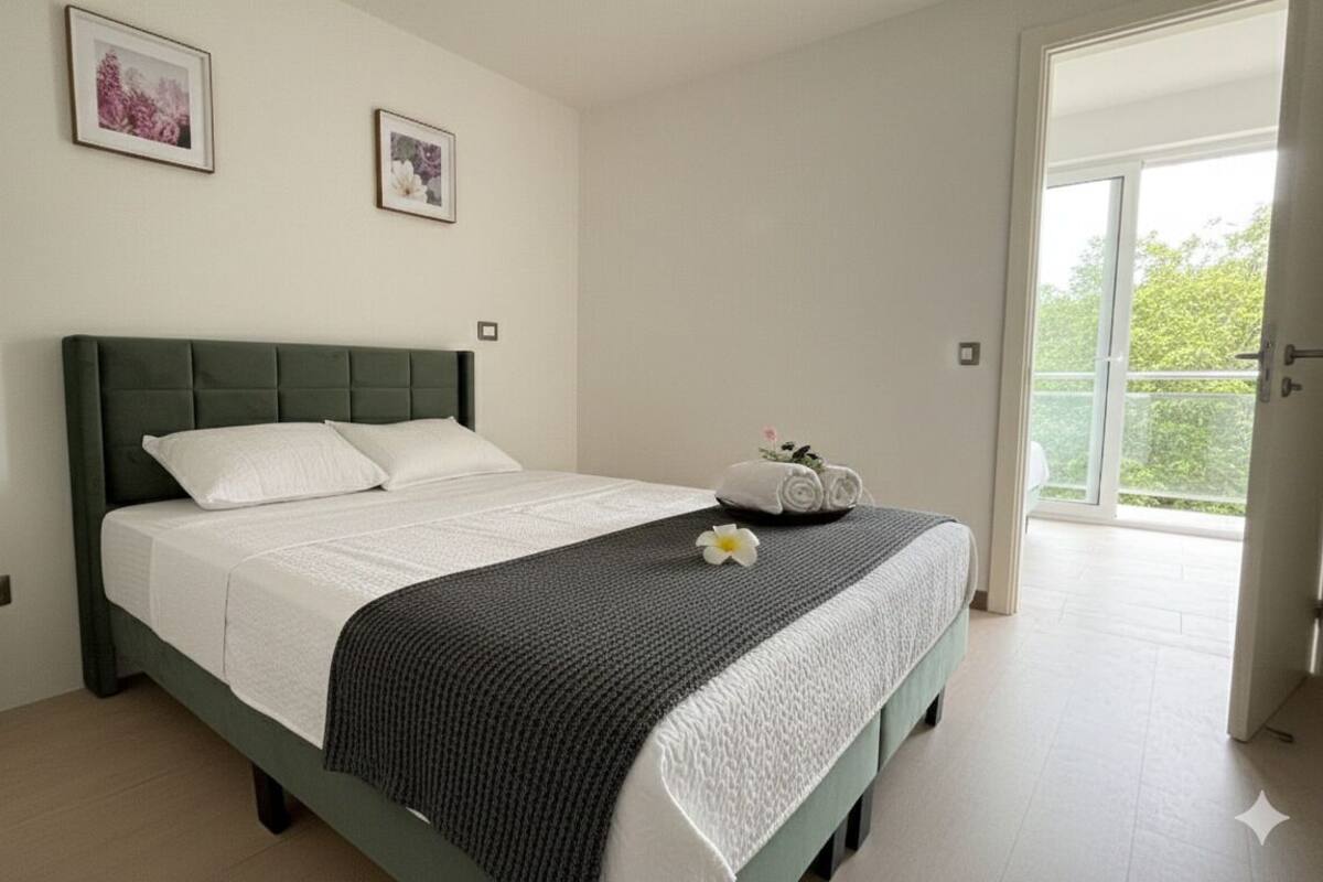 Ein Schlafzimmer mit einem grünen, gepolsterten Kopfteil, einer weißen Bettdecke und einer schwarz-weißen, strukturierten Tagesdecke. Auf dem Bett steht ein kleiner, weißer Vasen mit Blumen.