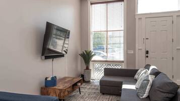 A living room with a grey sofa, a wooden coffee table, a TV mounted on the wall, and a large window with blinds.