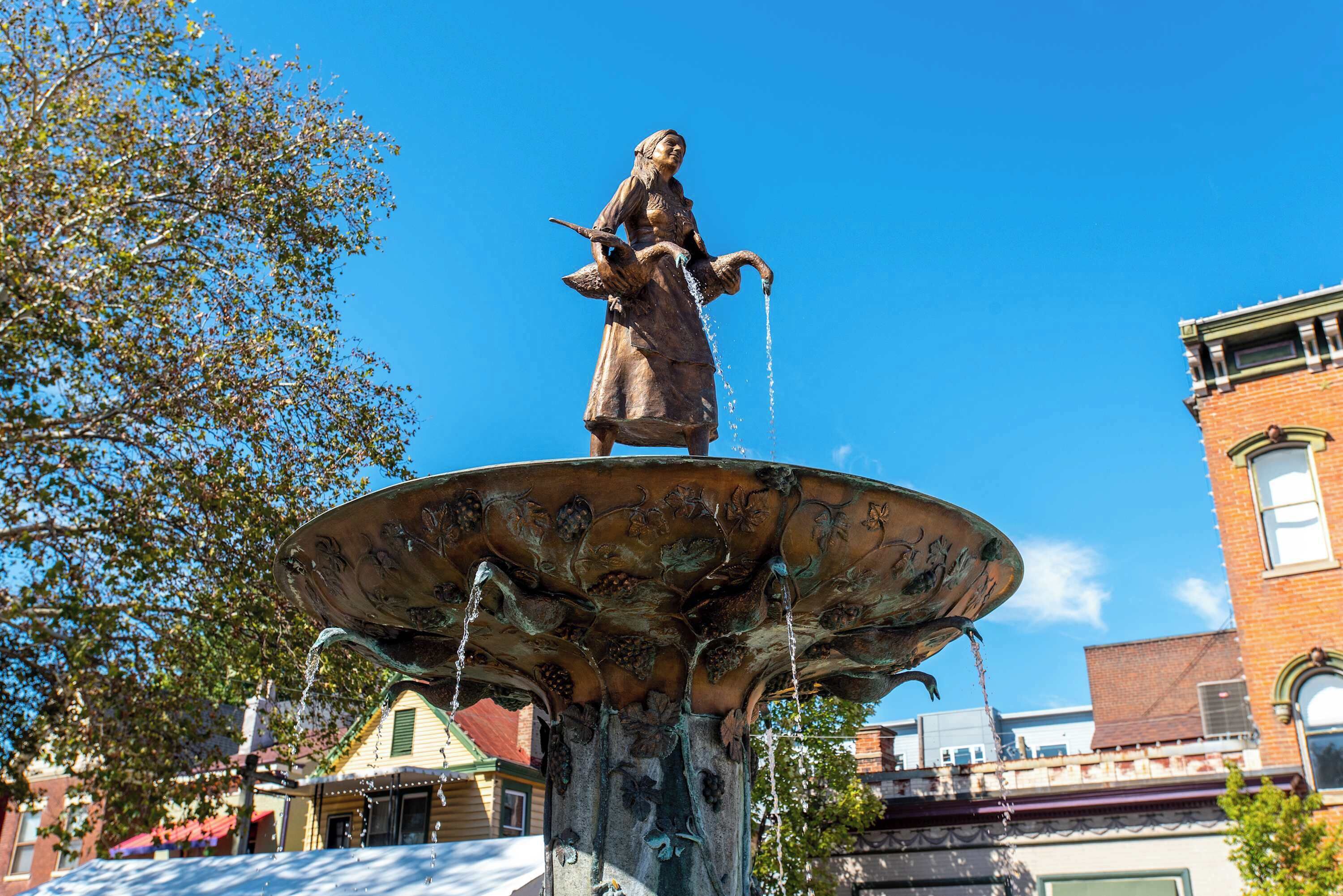Eine bronzene Statue einer Frau, die Wasser aus einem Krug gießt, steht auf einem verzierten Brunnenfundament, im Hintergrund sind Gebäude und Bäume zu sehen.