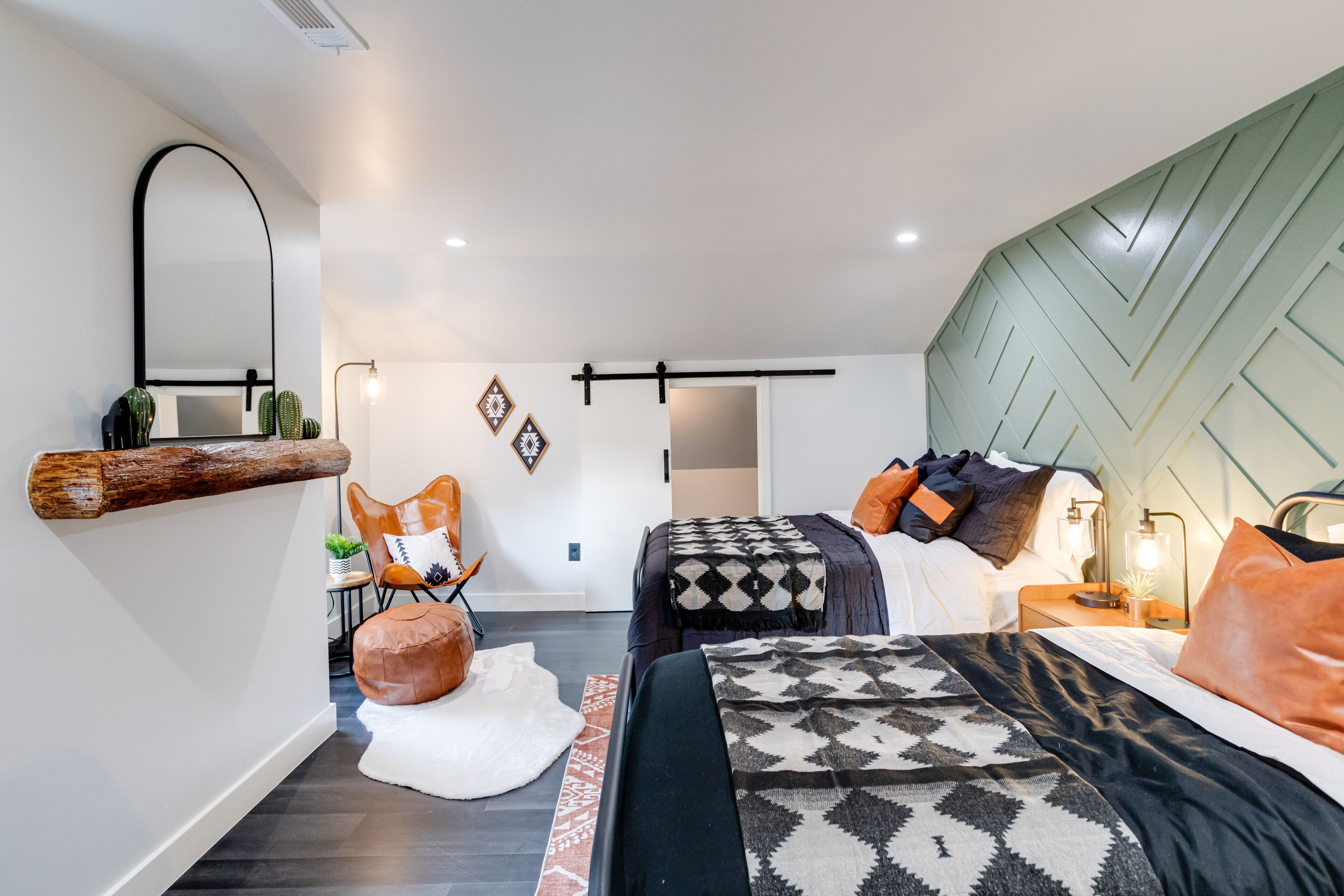 Een moderne slaapkamer met een groot bed, een kleiner bed, een bank en een stoel. Er hangt een spiegel aan de muur en op de vloer staat een plant.