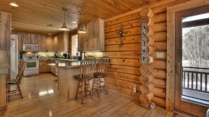 A log cabin interior with a kitchen area featuring wooden cabinets, a central island with bar stools, and a large window with a view of trees. - Beautiful Log Cabin with Acreage (Estes Park)