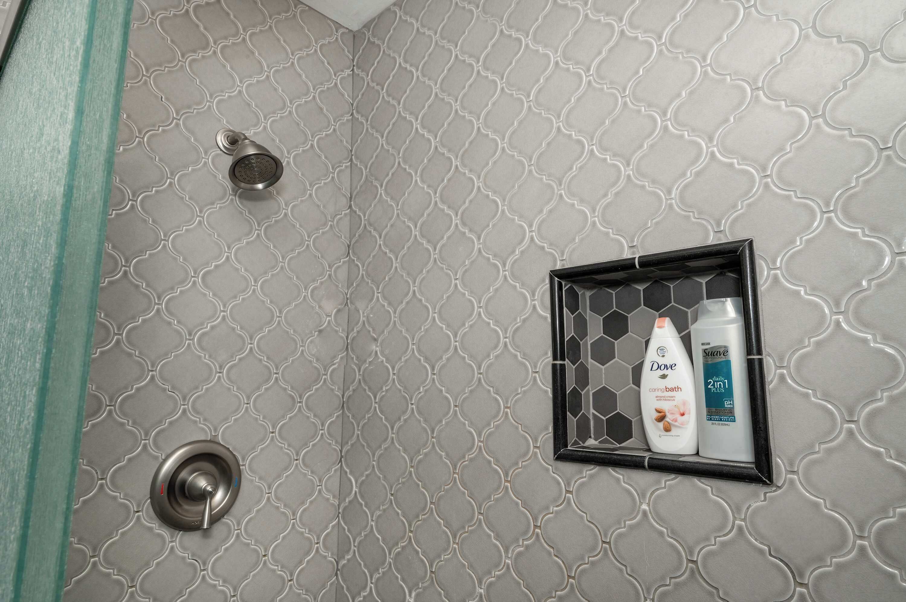 Een douche met een geometrisch betegeld patroon, een glazen deur en een zwart plankje met Dove en Suave producten.