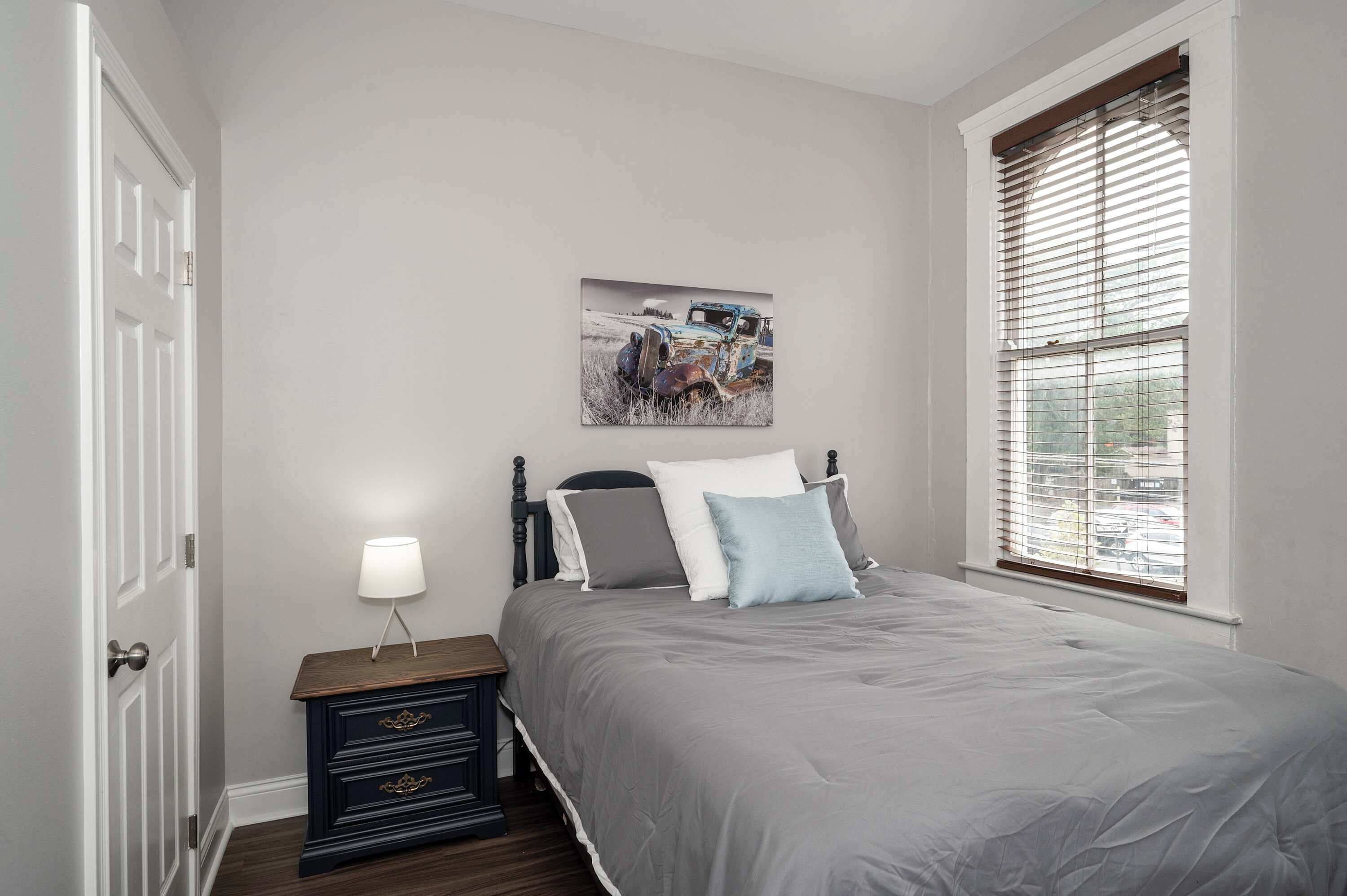 A bedroom with a bed, a nightstand, a lamp, a window with blinds, and a picture on the wall.