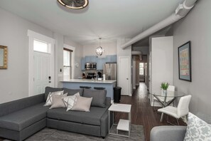 A modern living room with a grey sectional sofa, a glass coffee table, and a white armchair. A kitchen with blue cabinets and stainless steel appliances is visible in the background. - Heart of Mainstrasse 3 (Covington)