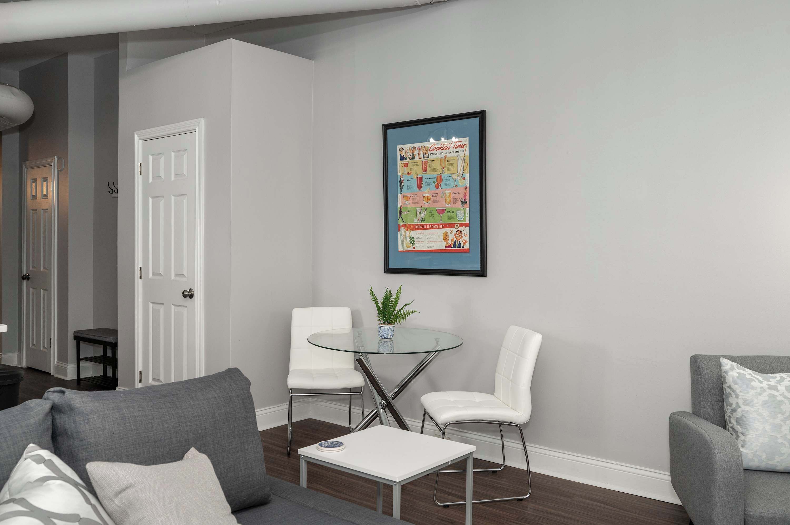 Una sala de estar moderna con una mesa de centro de vidrio, dos sillas blancas, un sofá gris y un cartel enmarcado en la pared.