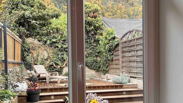 Blick durch eine Glastür auf eine Terrasse mit Holzboden, Gartenmöbeln und einer Blumenkomposition.