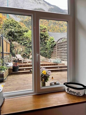 A view through a glass door of a patio area with a wooden deck, outdoor furniture, and a flower arrangement. - CYCLIST FRIENDLY HOUSE, CENTRE OF BOURG D’OISANS - SLEEPS 4 (Le Bourg-d'Oisans)