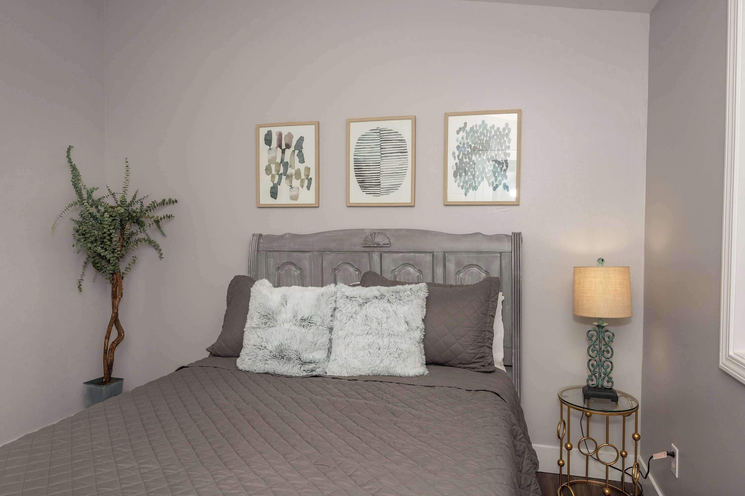 A bedroom with a bed, a nightstand with a lamp, and three framed artworks on the wall.