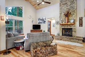 A cozy living room with a stone fireplace, a grey sofa, a television, and a large window overlooking trees.