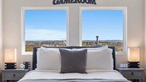 A bedroom with a bed, two bedside tables, a night lamp, a digital clock, and a view of a distant cityscape through two windows. - A Little Slice of Branson - Ozarks Views (Branson)
