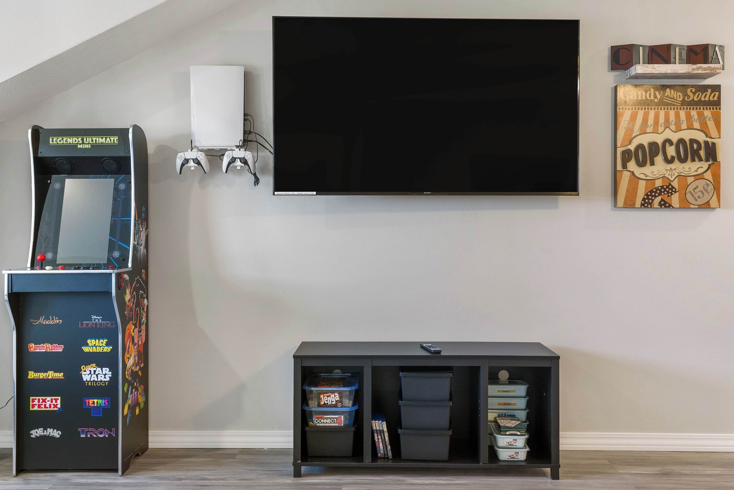 A room with a flat-screen TV mounted on the wall, a gaming console, and a shelf with storage boxes.