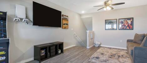 A living room with a flat-screen TV, gaming console, and a ceiling fan.