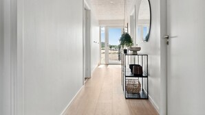Um corredor com piso de madeira, paredes brancas e uma porta que dá para o exterior. Há uma estante de metal preta com uma cesta trançada e um vaso de planta.
