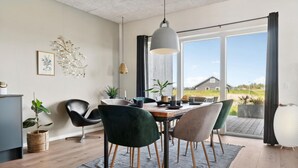 Uma sala de jantar com uma mesa de madeira, quatro cadeiras, um lustre e vista para uma casa através de grandes janelas.