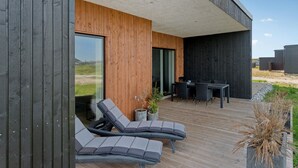 Um deck moderno ao ar livre com cadeiras de descanso e uma mesa de jantar, cercado por paredes de madeira e grandes janelas.