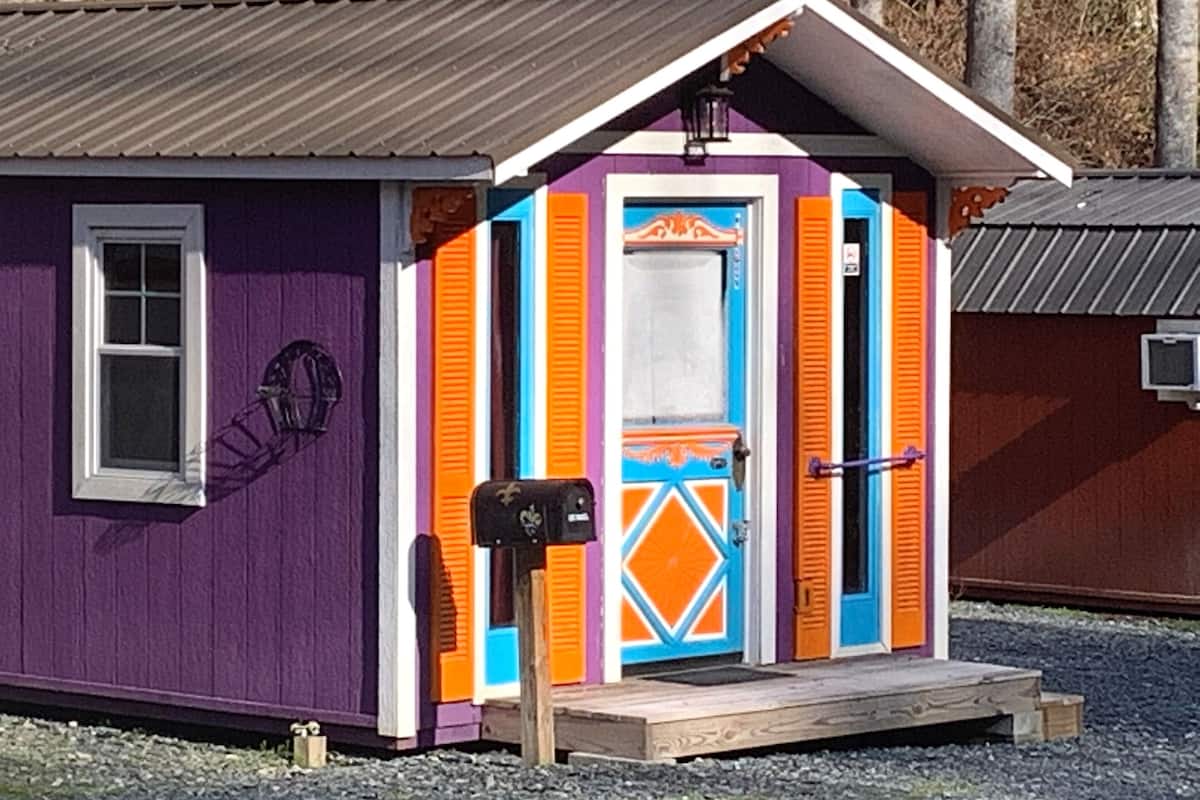 Pieni, värikäs varasto, jonka ulkoseinät ovat violetit, ovi valkoinen ja ikkunaluukut oranssit.