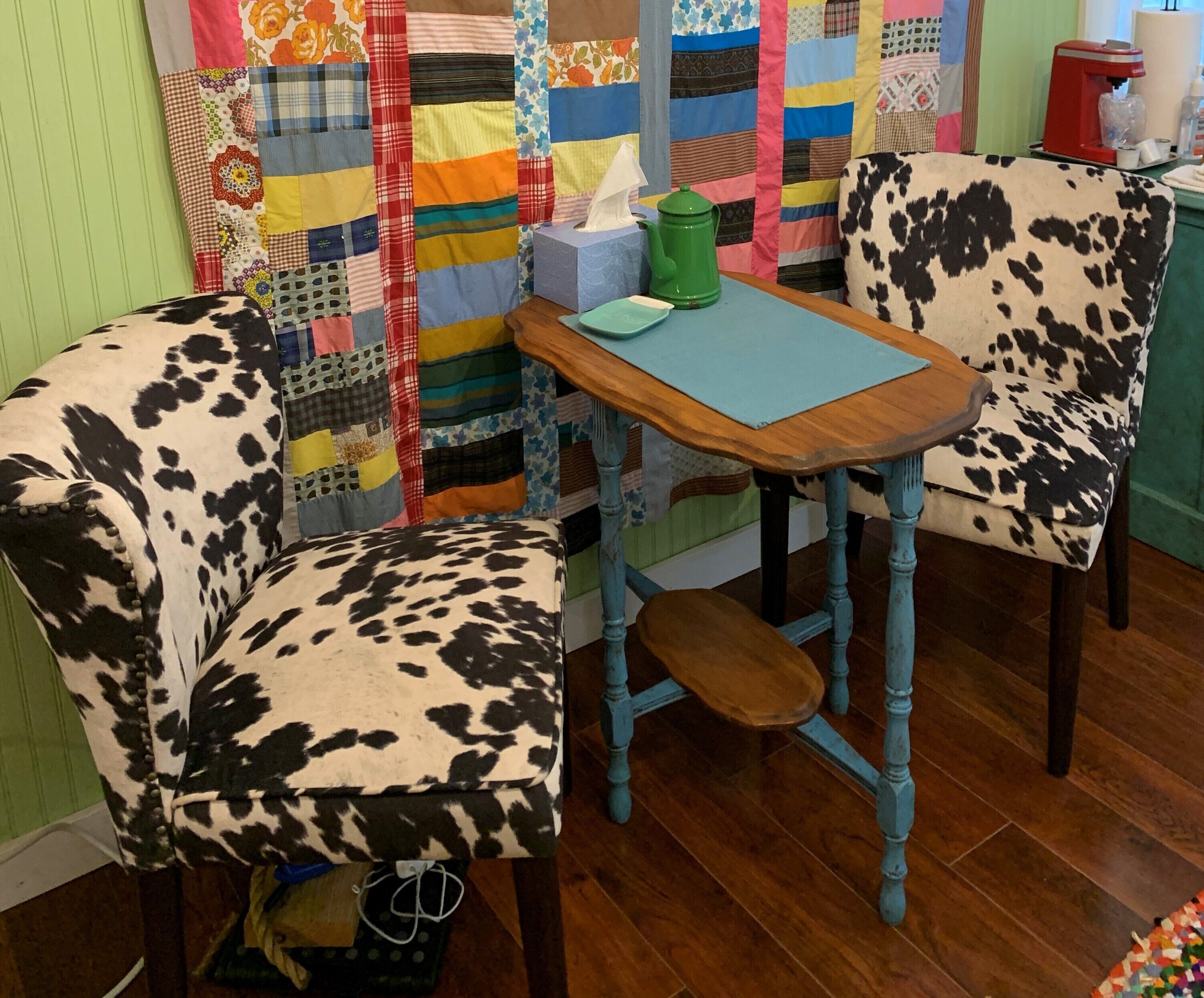 Un pequeño comedor con una mesa de madera y dos sillas con estampado de cuero. Hay una caja de pañuelos y una cafetera verde sobre la mesa.