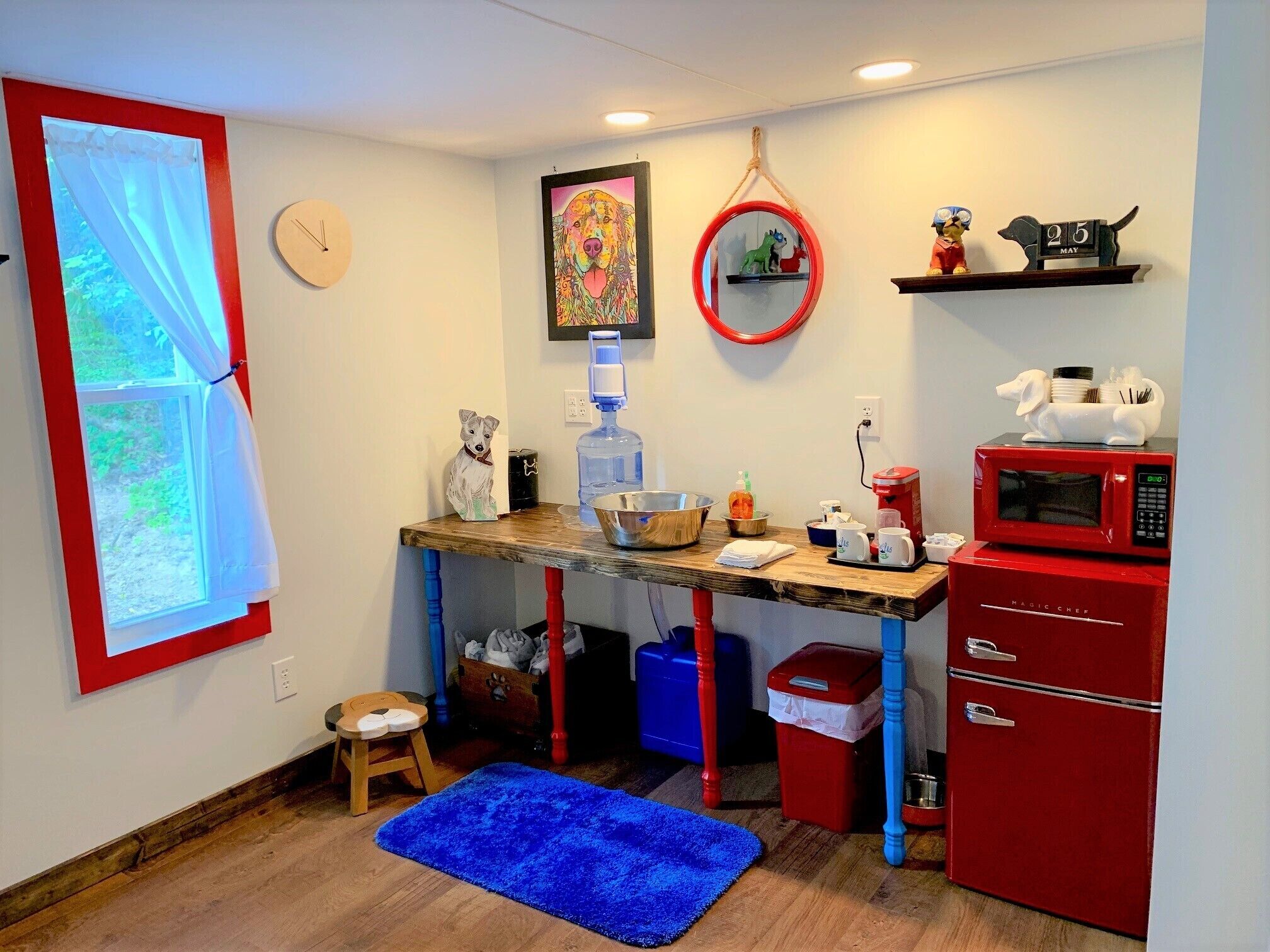A compact kitchen with a wooden table, a red refrigerator, a microwave, a sink, and a window with curtains.
