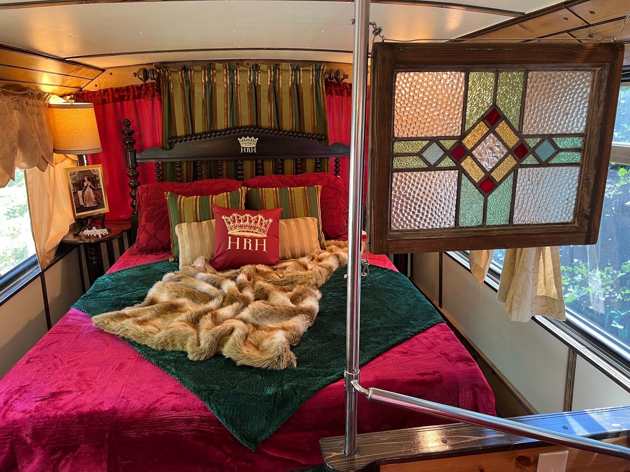 Ein Schlafzimmer mit einem Bett, einem Kopfteil mit einem Kronenwappen, einem roten Kopfkissen mit dem Schriftzug "HRH" und einem Fenster mit geometrischem Muster.