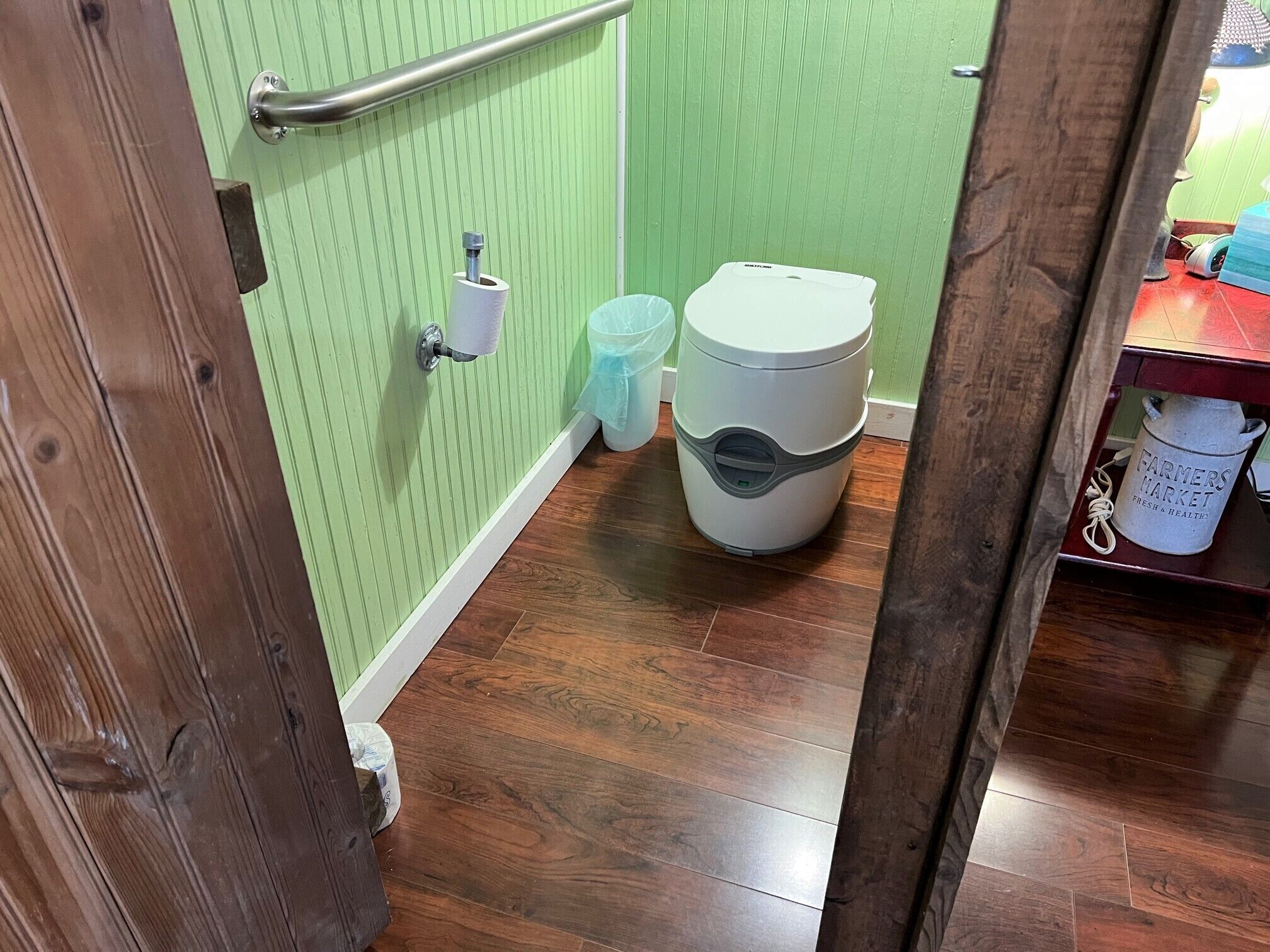 A bathroom with a green wall, a wooden floor, a toilet, a trash bin, and a metal bucket labeled "Farmers Market Fresh & Healthy."