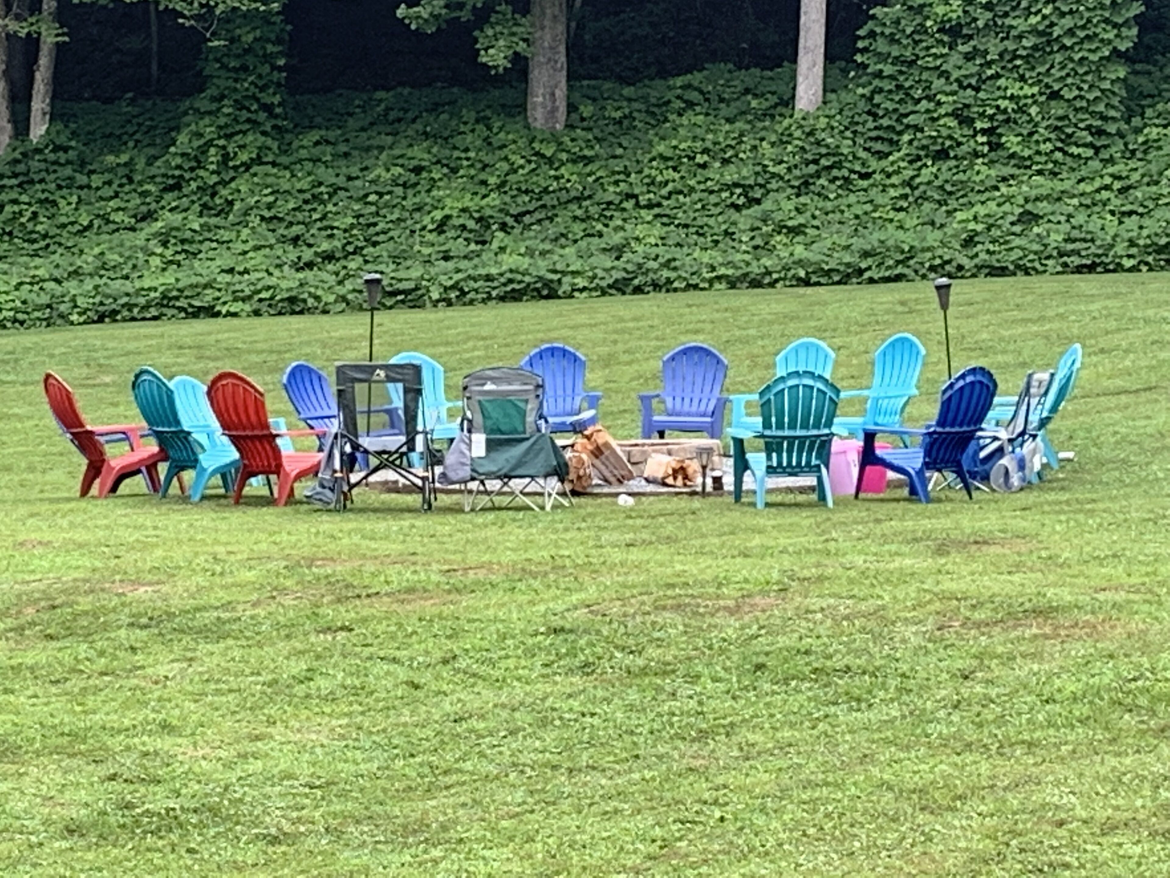 A row of colorful Adirondack chairs arranged in a circle on a grassy field with a dense hedge in the background.