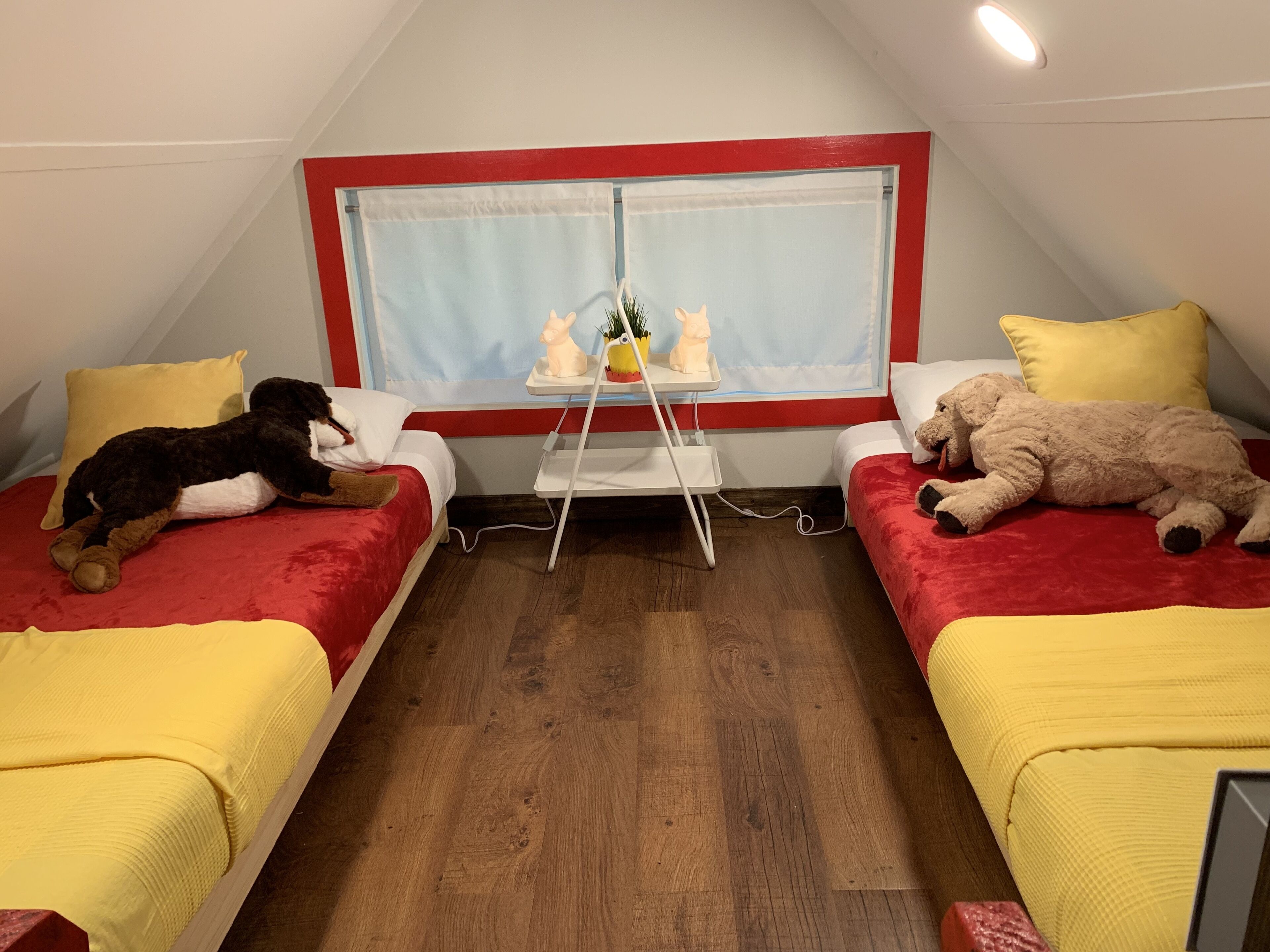 Une petite pièce chaleureuse, dotée de deux lits, chacun recouvert d’une couvre-lit jaune et agrémenté d’une tête de lit rouge. On y trouve une petite table sur laquelle sont posées une plante et deux statuettes.