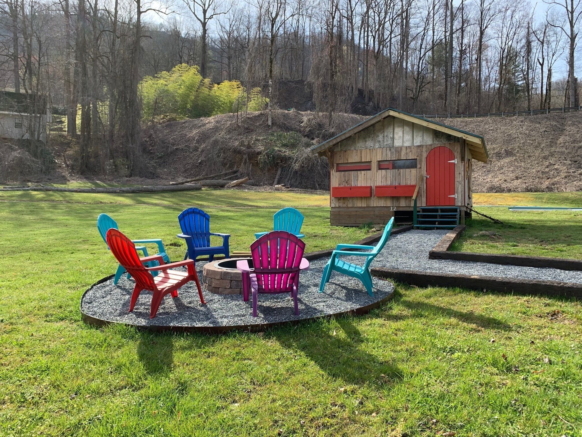 Eine Holzhütte mit roter Tür, umgeben von einer grünen Wiese mit bunten Adirondack-Stühlen und einer Feuerstelle.