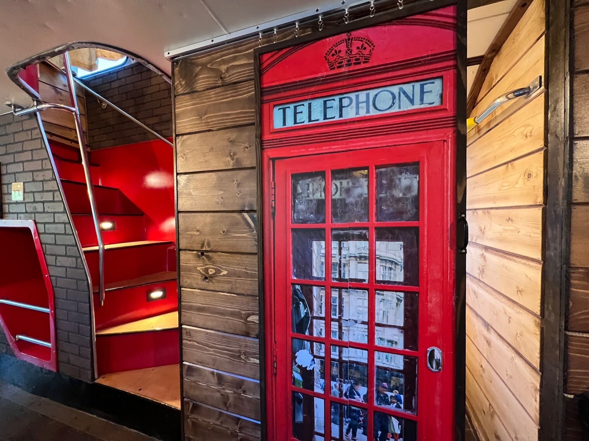 A red telephone booth with a crown emblem, a staircase, and a wooden wall with a window reflecting a busy street scene.
