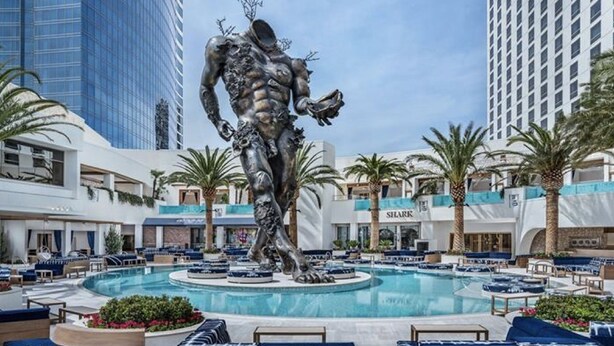 Zona de piscina con una escultura central, rodeada de palmeras y edificios modernos.