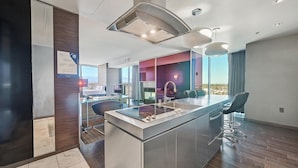 Habitación de hotel moderna con una isla grande, un área de comedor separada con taburetes de bar, un televisor de pantalla plana y vistas a la ciudad.