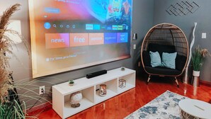 Una sala de estar moderna con un televisor de pantalla plana grande mostrando un canal de videojuegos, un sillón de mimbre, una consola blanca y un globo decorativo.
