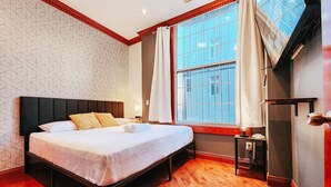 Una cama bien hecha con almohadas, una mesita de noche con lámpara, y un amplio ventanal con cortinas.