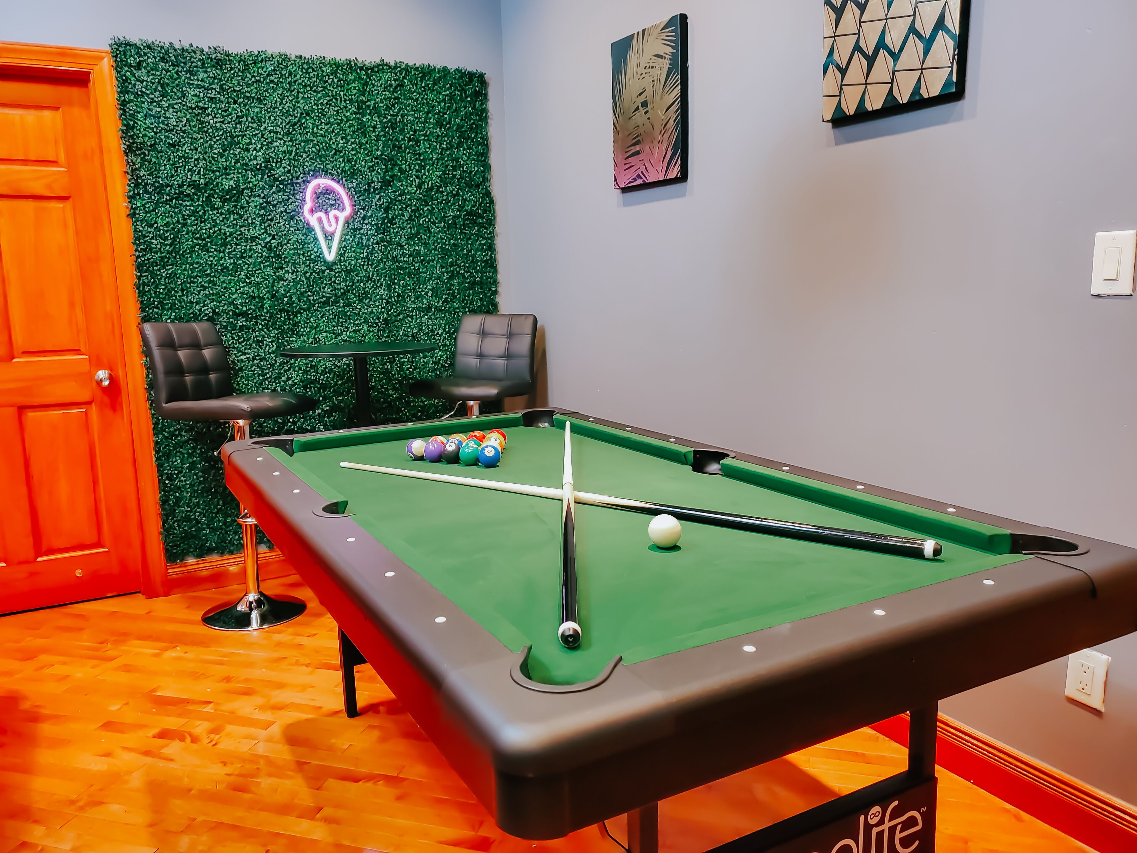 Una mesa de billar con bolas, dos sillas, un letrero de neón y una pared verde.