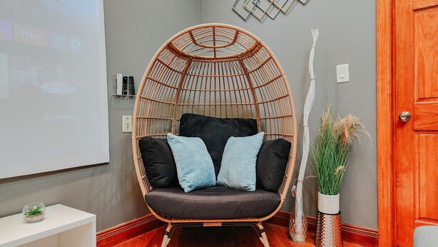 Una habitación con una silla de mimbre, una mesa consola blanca, una planta en maceta y una puerta de madera.