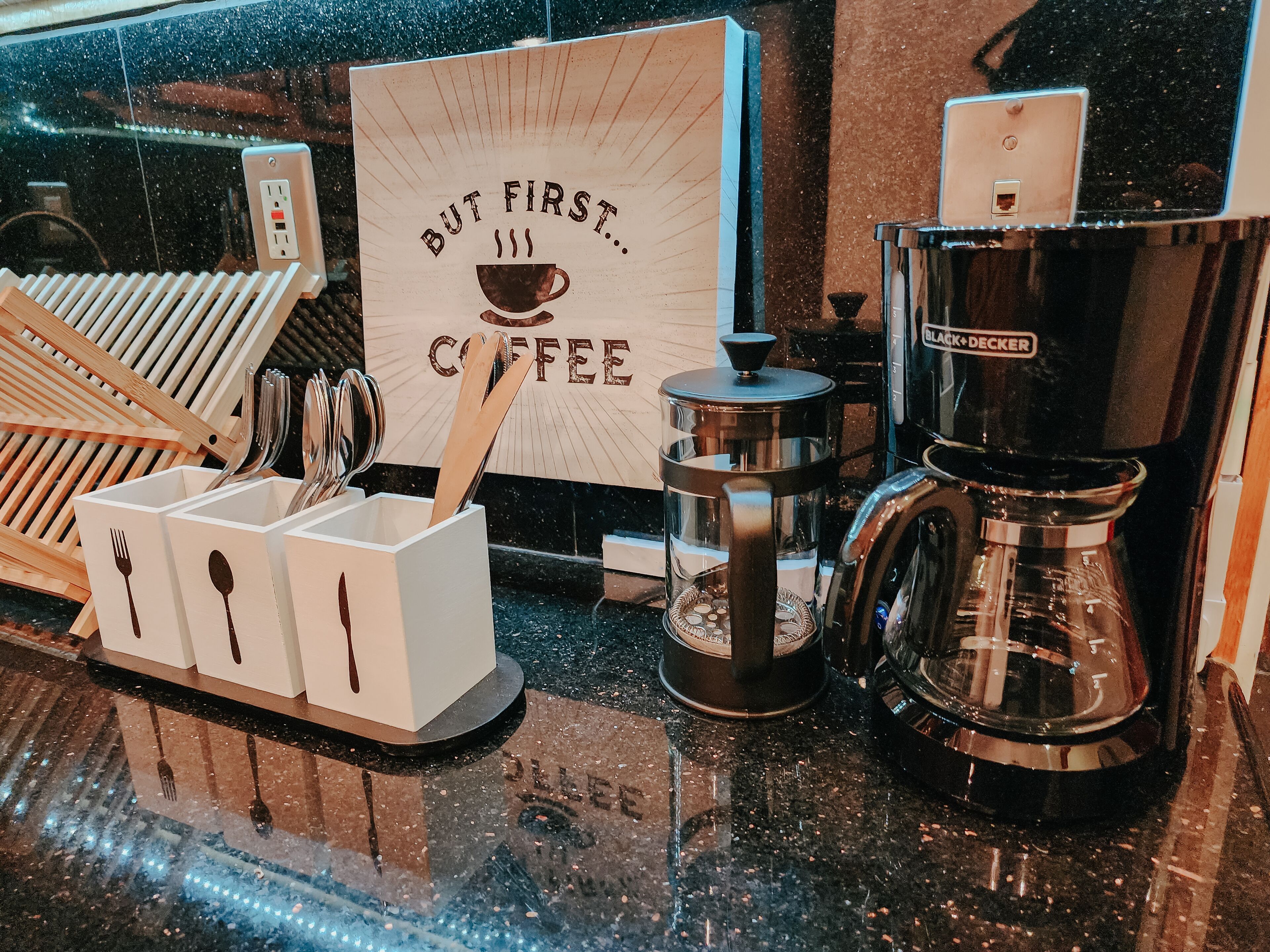 Una estación de café con un letrero que dice “But First… Coffee”, una cafetera y utensilios.