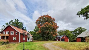 A red barn with a white door, a wooden porch, and a gravel driveway leading to a large tree with orange leaves. - Beautiful home in älmhult with kitchen (Älmhult)