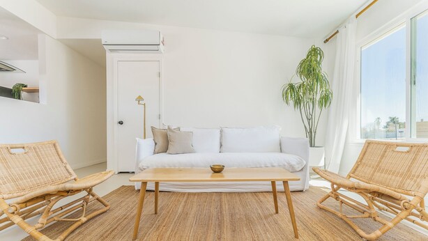 Un salon moderne avec un canapé blanc, deux fauteuils en osier, une table basse en bois et une grande fenêtre donnant sur un beau paysage.