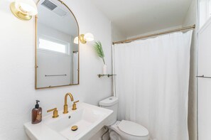 A bathroom with a white sink, gold faucets, a white toilet, a shower curtain, and a mirror with lights.