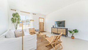 Un salon spacieux avec un canapé blanc, une table à manger en bois et un fauteuil en osier. Une grande fenêtre laisse entrer la lumière naturelle, et un téléviseur est fixé au mur.