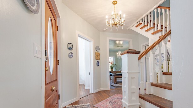 Un pasillo con puerta de madera, un candelabro y una escalera con barandillas blancas y escalones de madera.