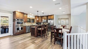 A modern kitchen with wooden cabinets, stainless steel appliances, and a dining area with wooden chairs and a white railing. - Bear Hollow Heaven Private Hot Tub 4 Bedroom Luxury Condo (Park City)