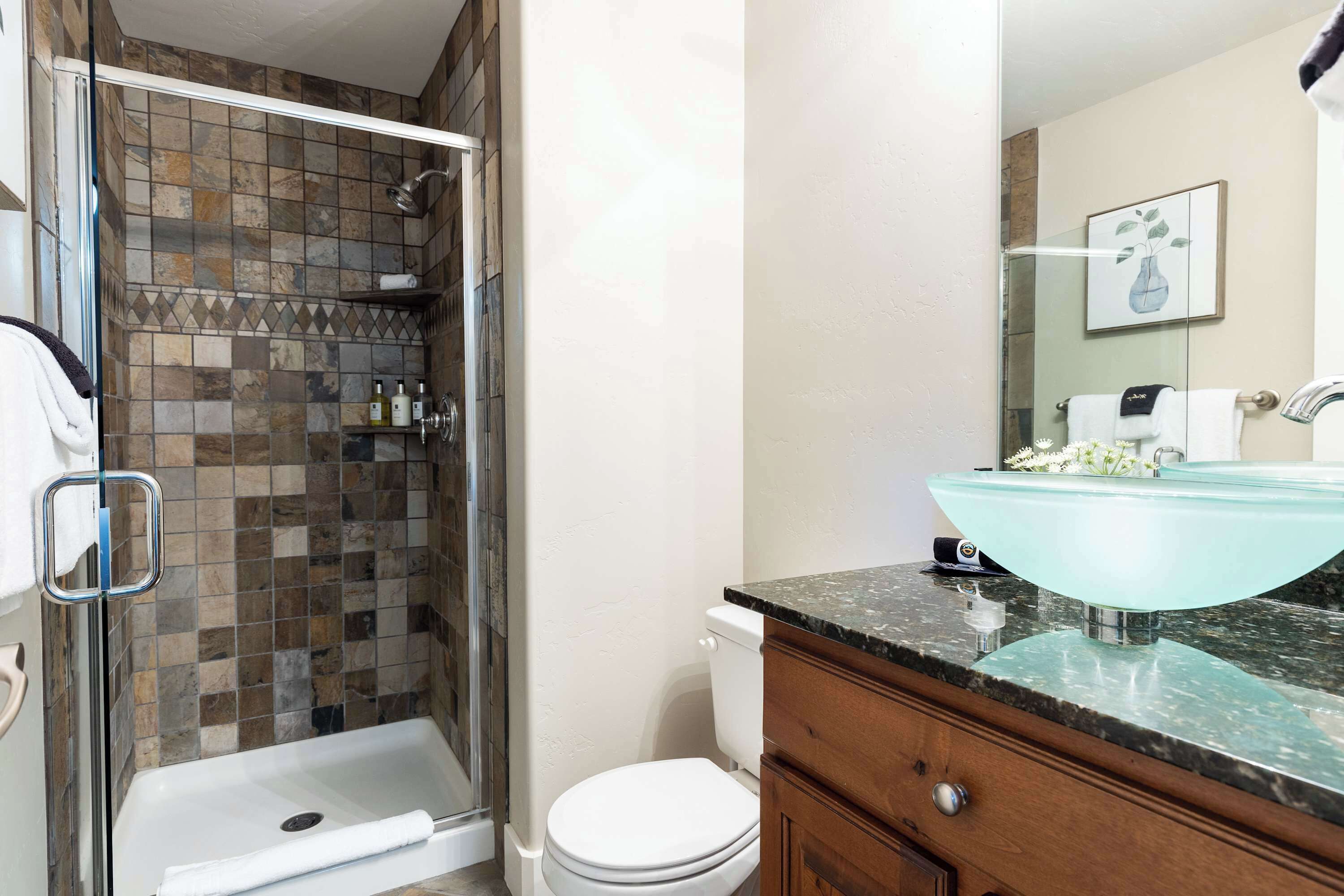 A bathroom with a glass shower enclosure, a white toilet, a marble sink, and a wooden vanity.